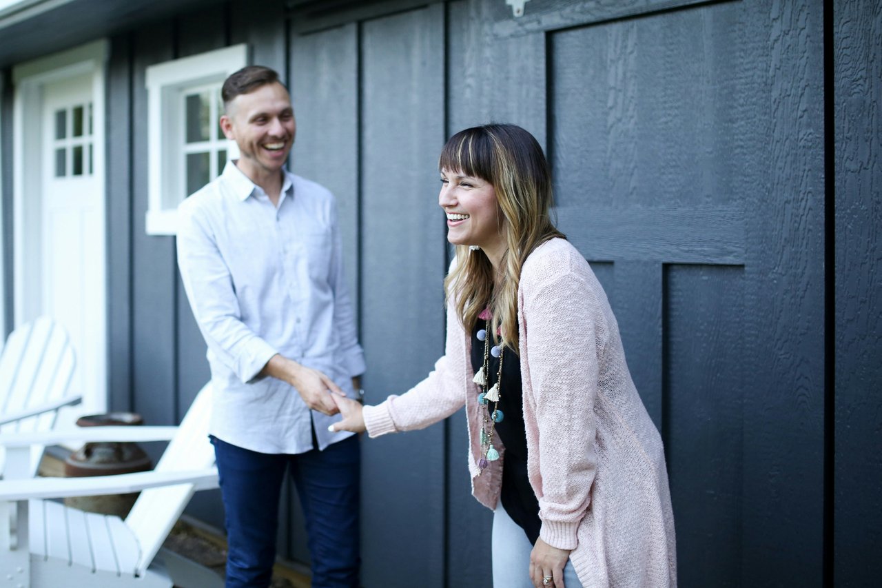 A smiling woman and man shake hands outside a dark-sided house.