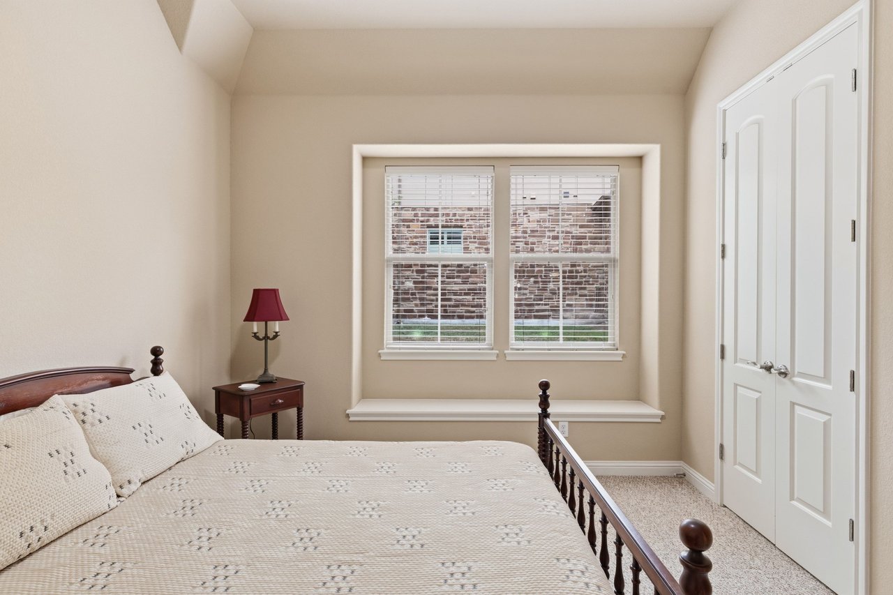 Bedroom with cream patterned bedding, a wooden bed, and a window seat beneath two windows.