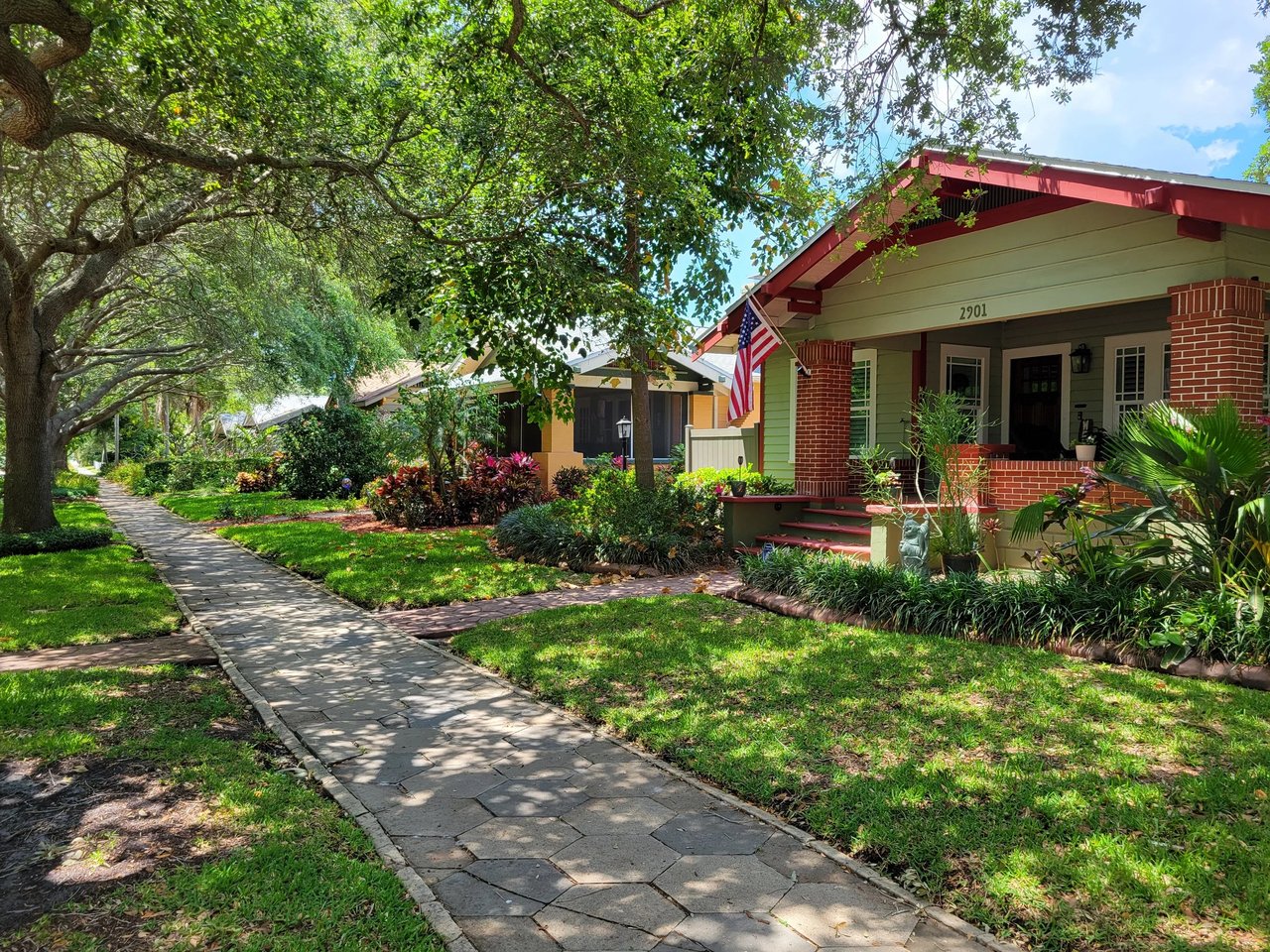 Bungalow with a red-trimmed porch and American flag beneath mature trees beside a paved sidewalk.