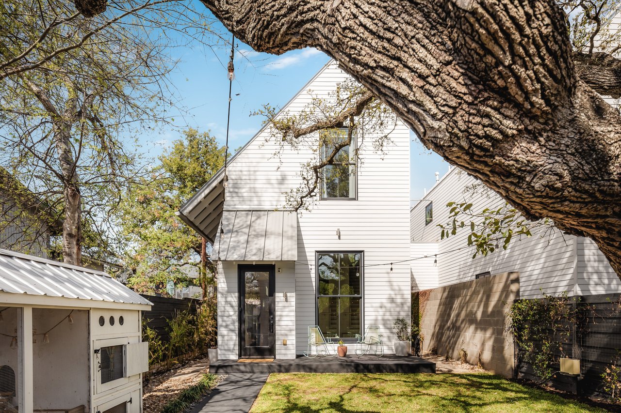 Modern white two-story home framed by a massive oak branch, with a small patio and backyard lawn.