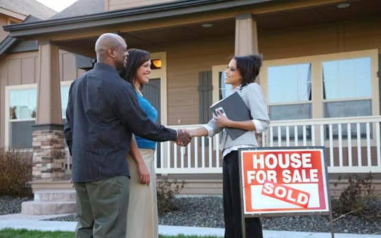 A couple shakes hands with an agent beside a sold sign outside a house.