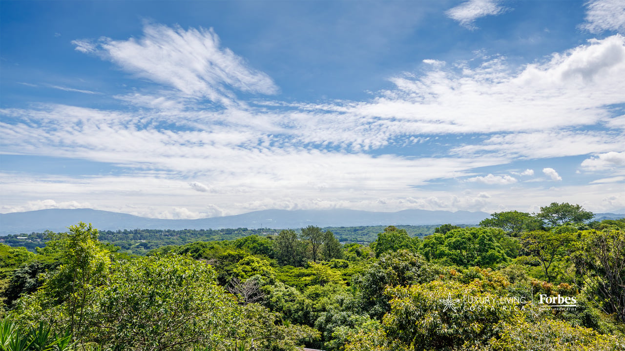 Green agricultural property with dense tropical greenery and distant mountain views beneath a cloud-filled sky.