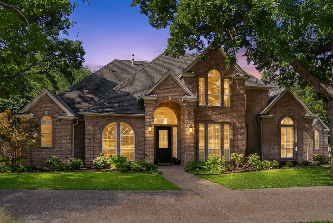 Two-story brick home at dusk, framed by mature trees, with glowing arched windows and a curved front walkway.