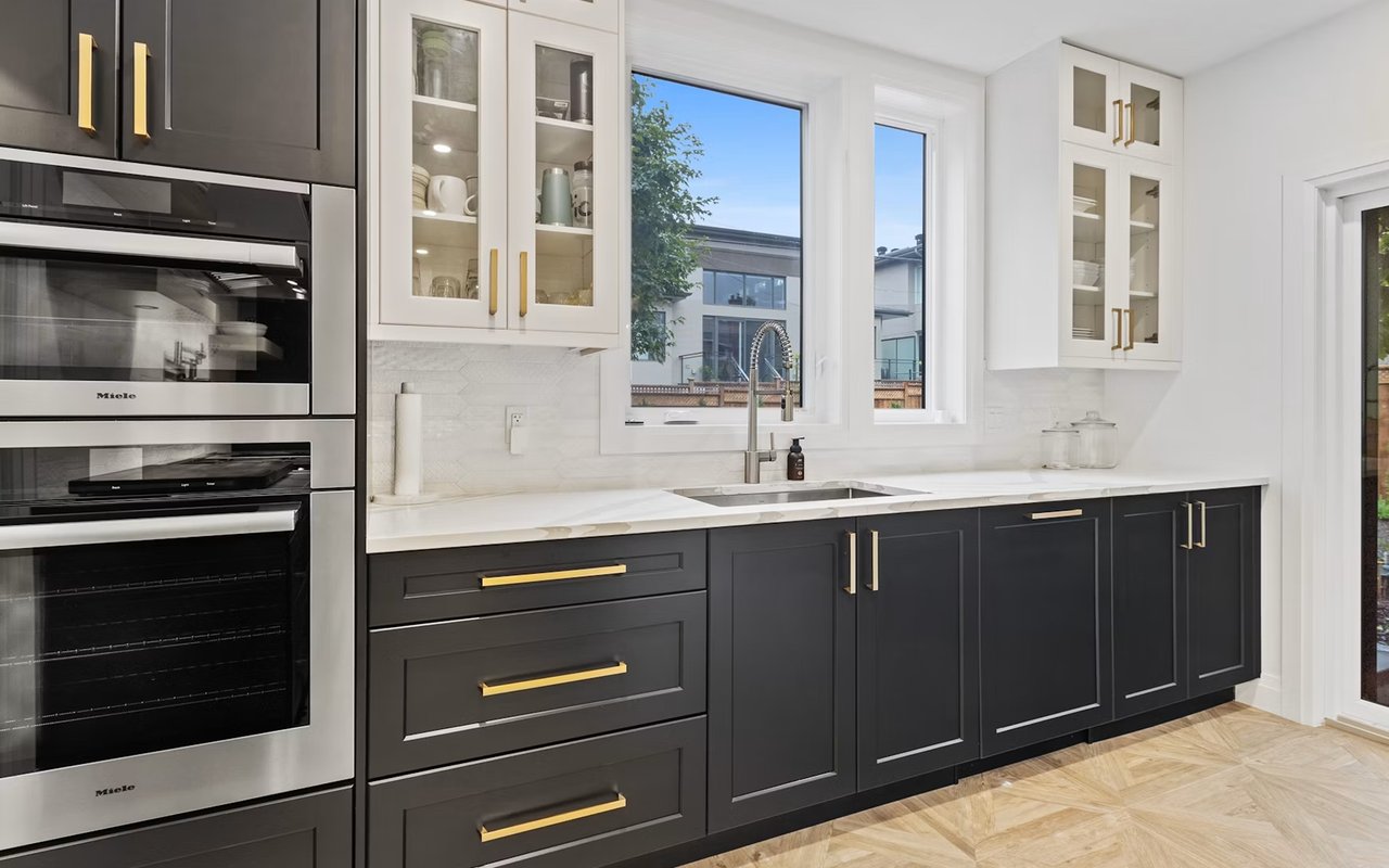 Modern kitchen with charcoal cabinetry, gold pulls, white counters, and stacked stainless-steel ovens.