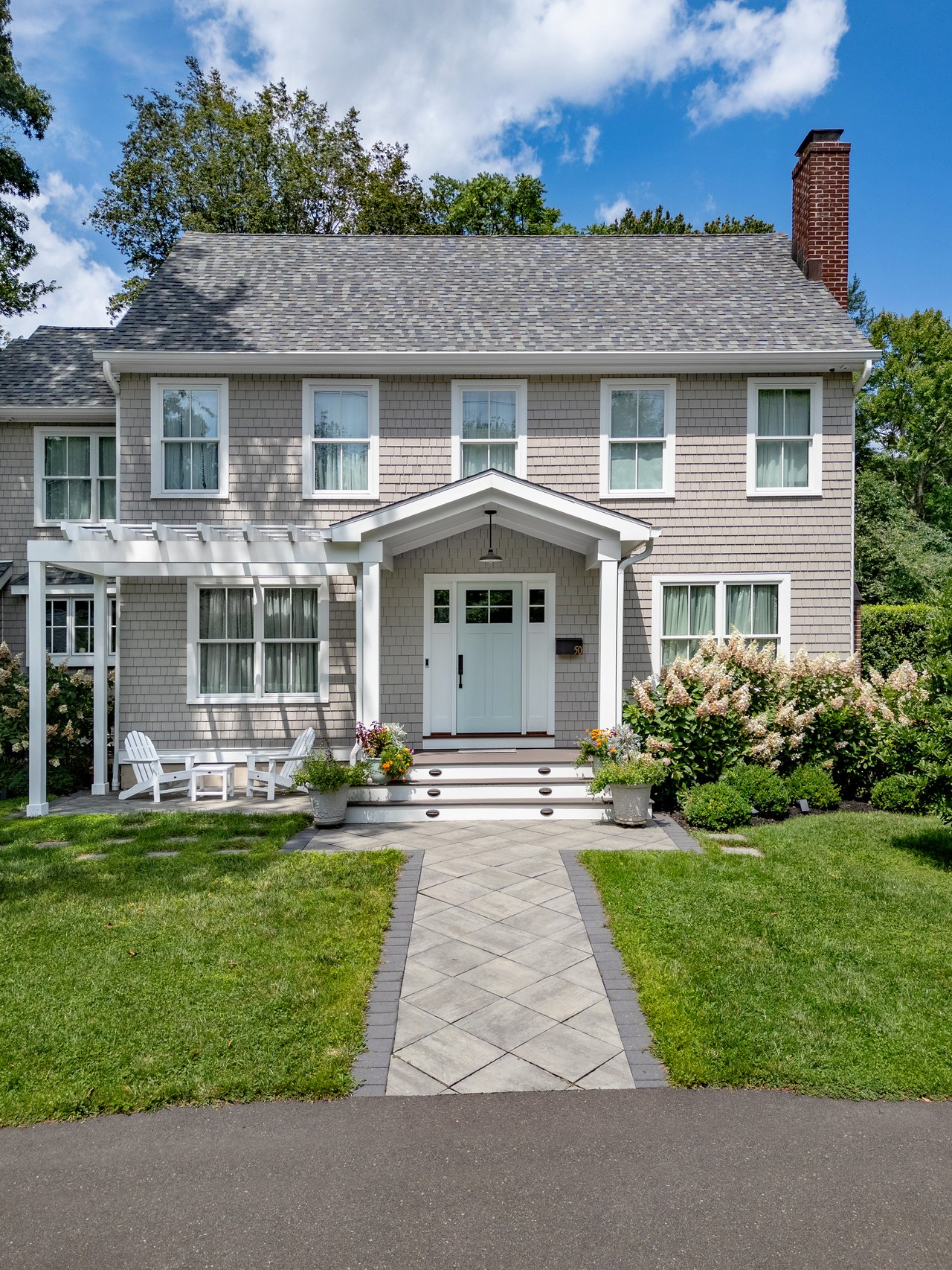 Gray-shingled Colonial with a brick chimney, white pergola, and paved front walkway.