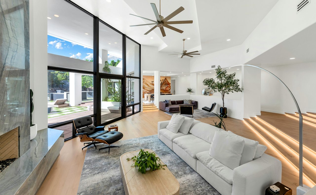 Modern living room with a white sectional, marble fireplace, tall glass windows, and a sculptural staircase.