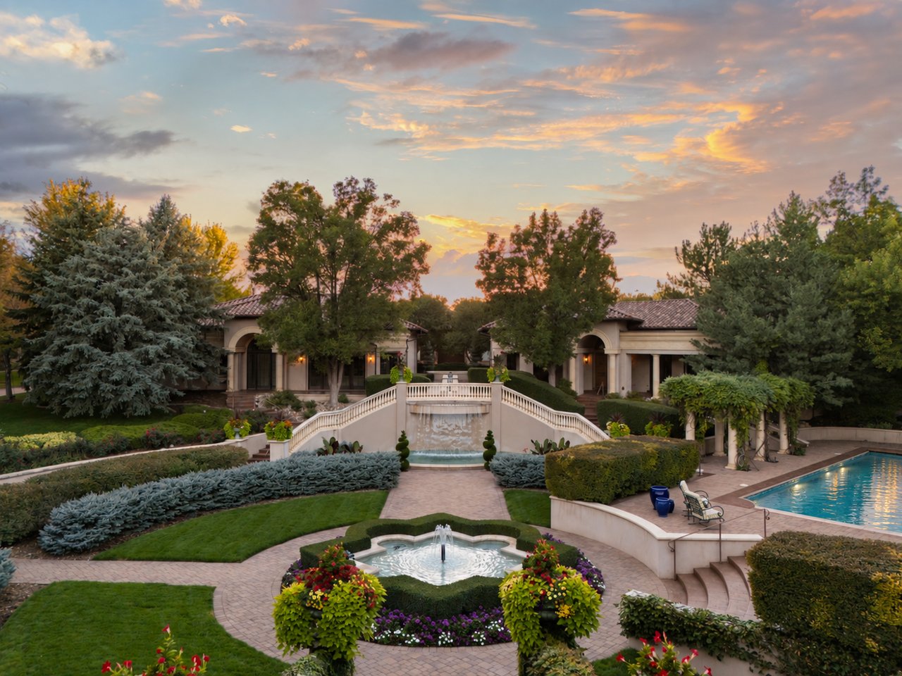 Estate gardens with a cascading fountain, colorful flower beds, mature trees, and a swimming pool at sunset.