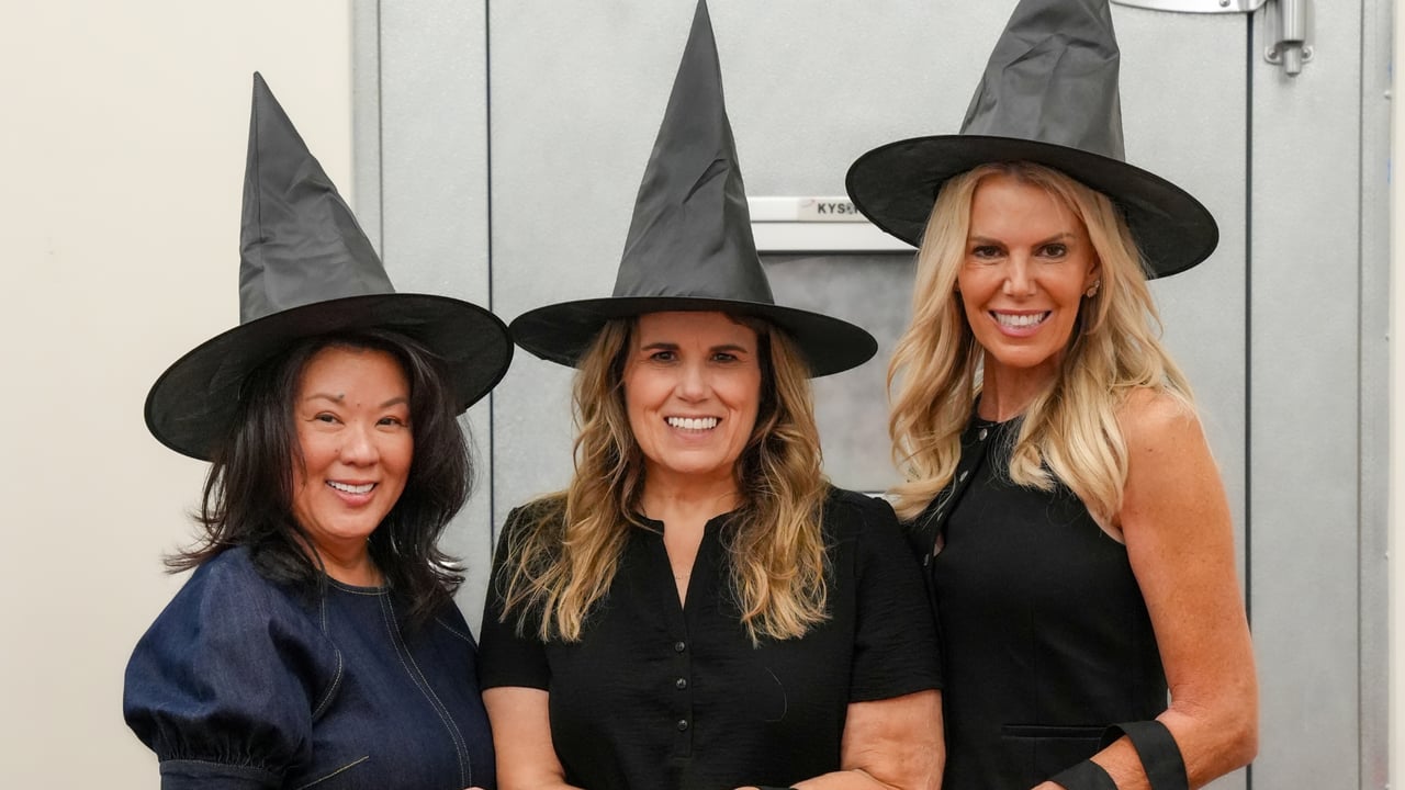 Three women smile together in witch hats.