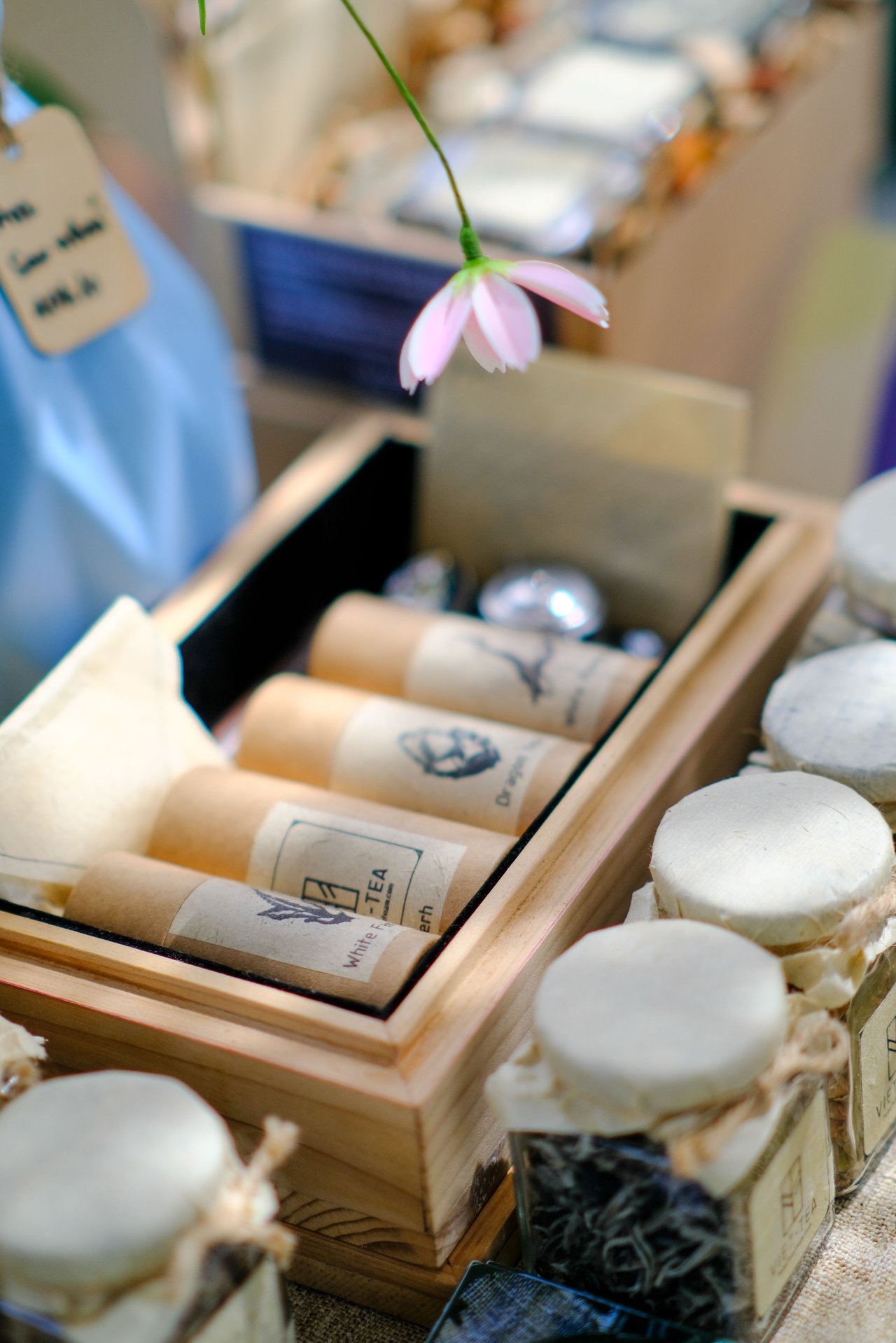 Wrapped tea products and round white soaps in wooden boxes beneath a small pink flower.