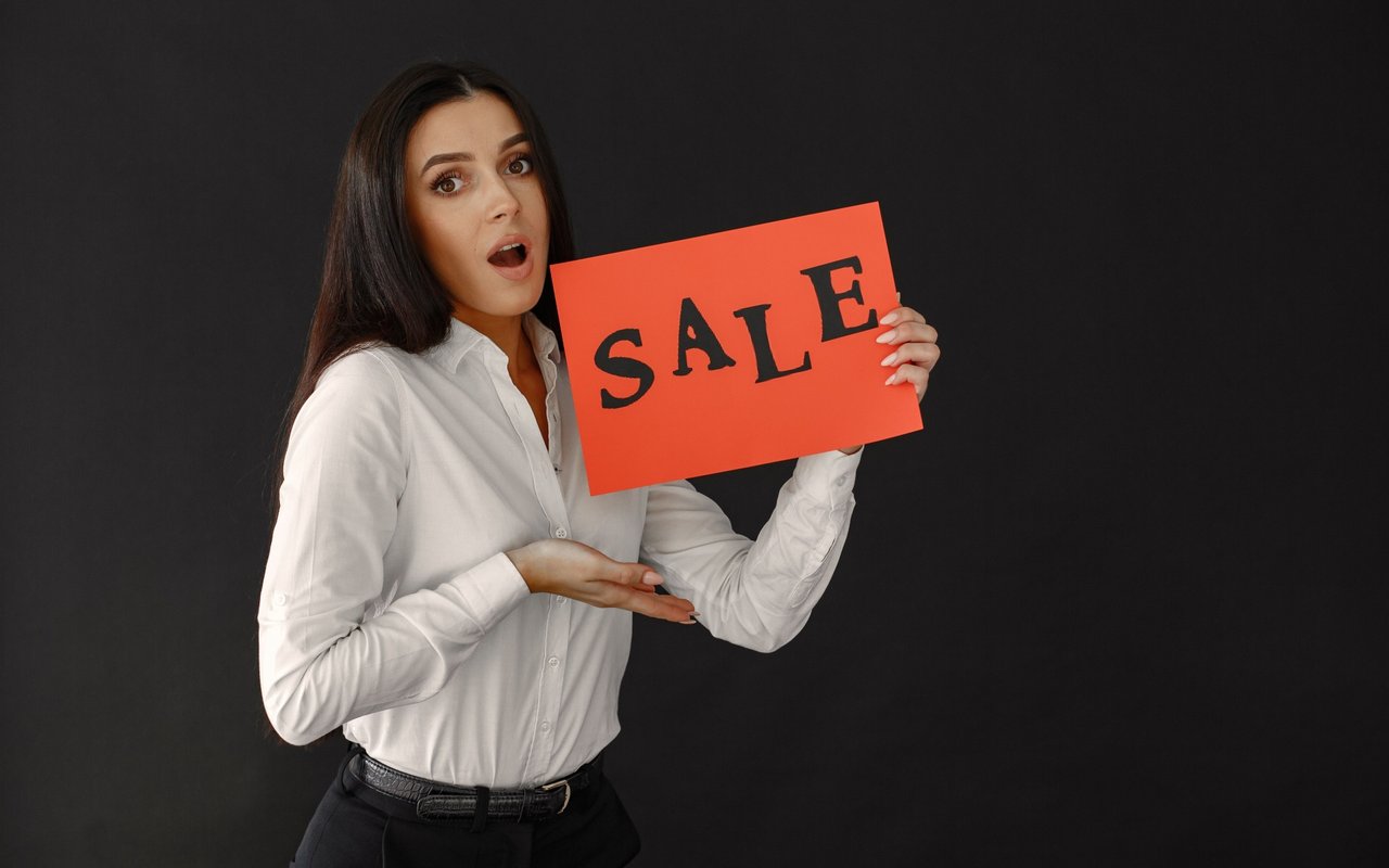 A woman holds a red sign reading “SALE” against a dark background.