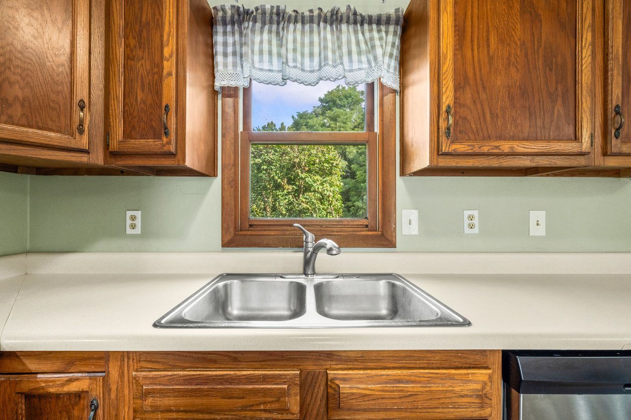 Kitchen sink beneath a window, framed by warm wood cabinets and a checkered valance.