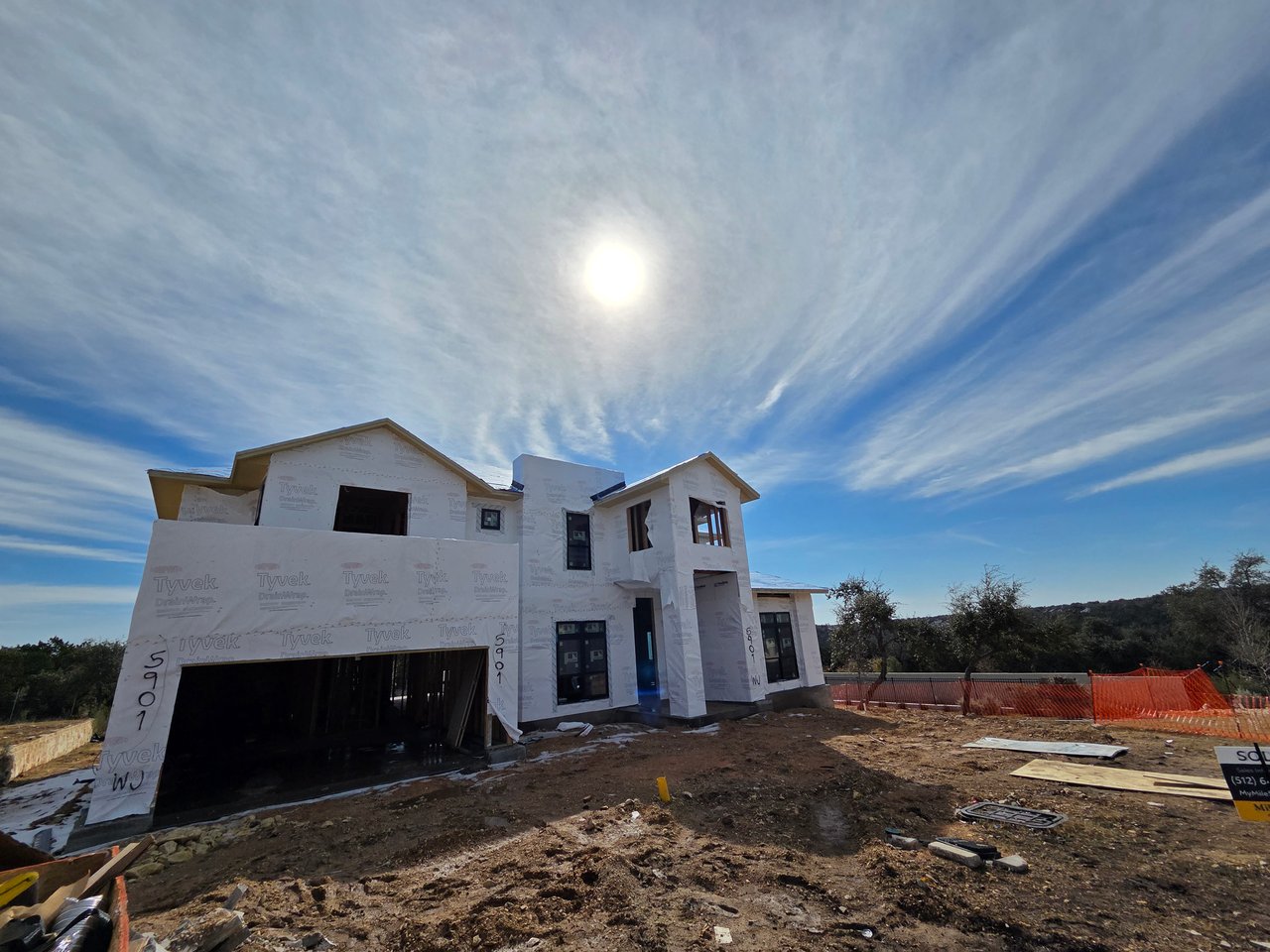 Two-story home under construction with Tyvek wrap, an open garage, and a dirt yard.