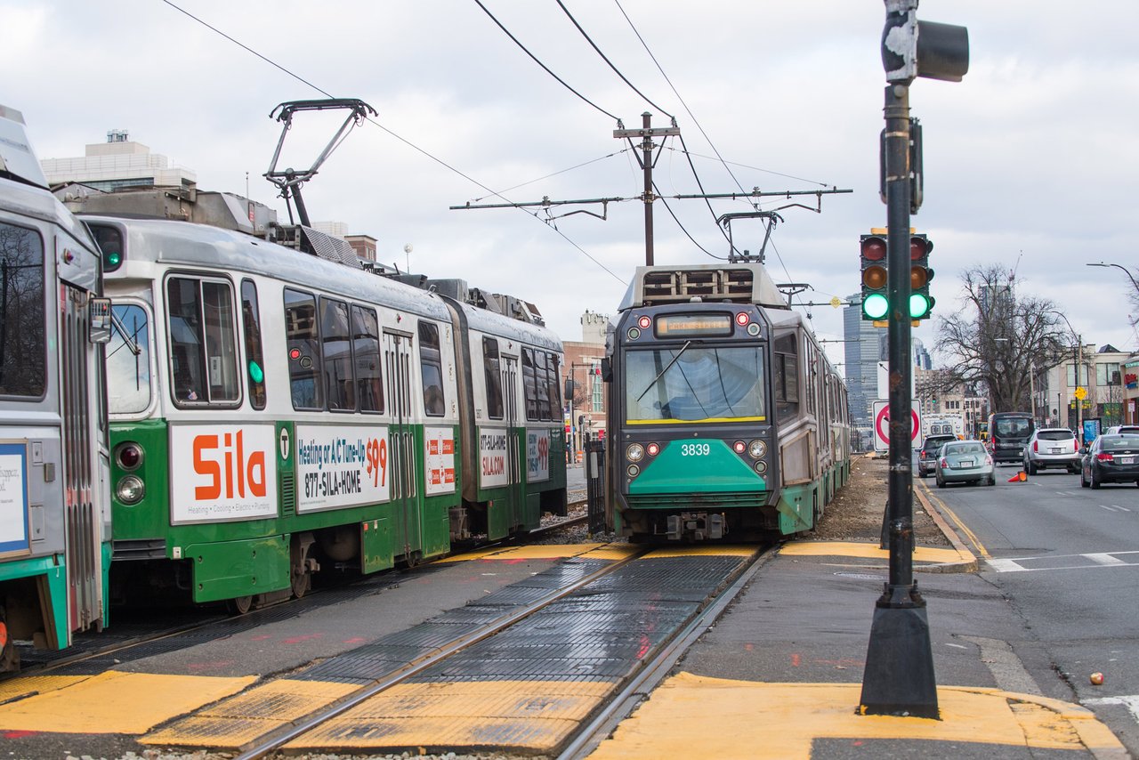 Two green-and-white light-rail trains crossing a city street beneath overhead wires.
