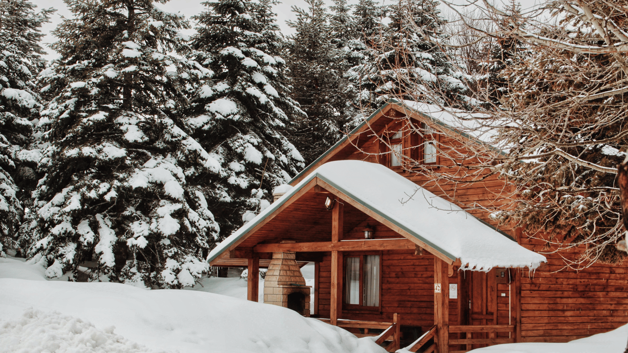 Wooden cabin with deep snow on its roofs, surrounded by snow-covered evergreens.