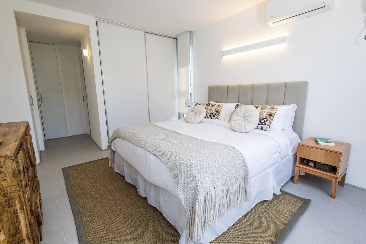 Bright bedroom with a white bed, tall upholstered headboard, patterned pillows, and sliding closet doors.
