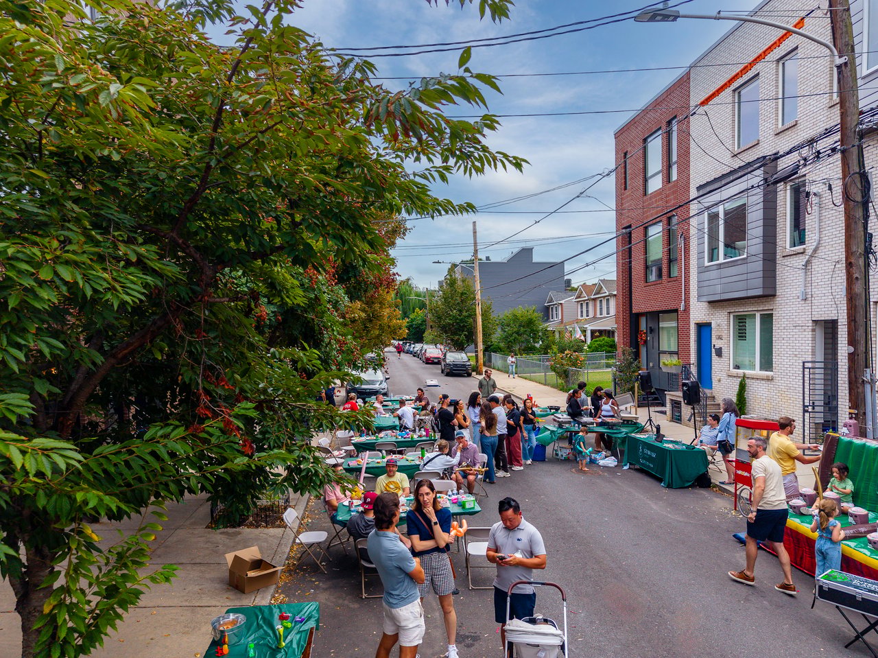 Neighbors gather at a street block party with picnic tables and food stands beside brick rowhouses.