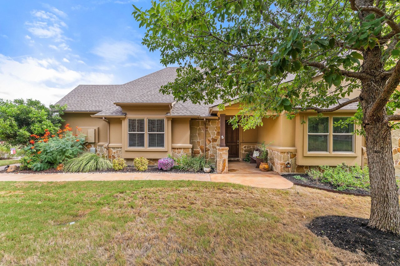 Single-story tan home with stone accents and a covered entry beneath a large leafy tree.