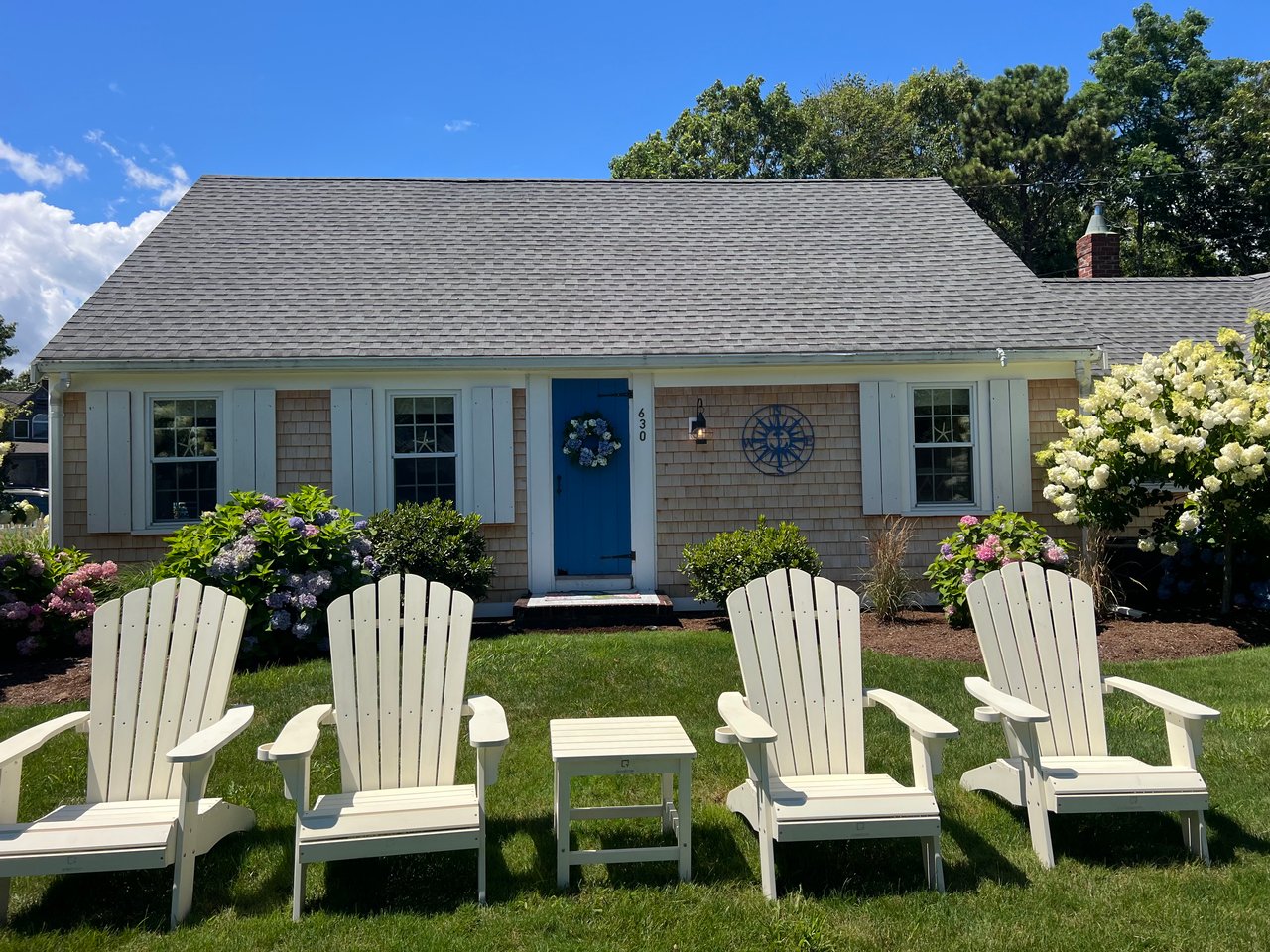 Shingle-sided cottage with a blue front door and white Adirondack chairs on the lawn.