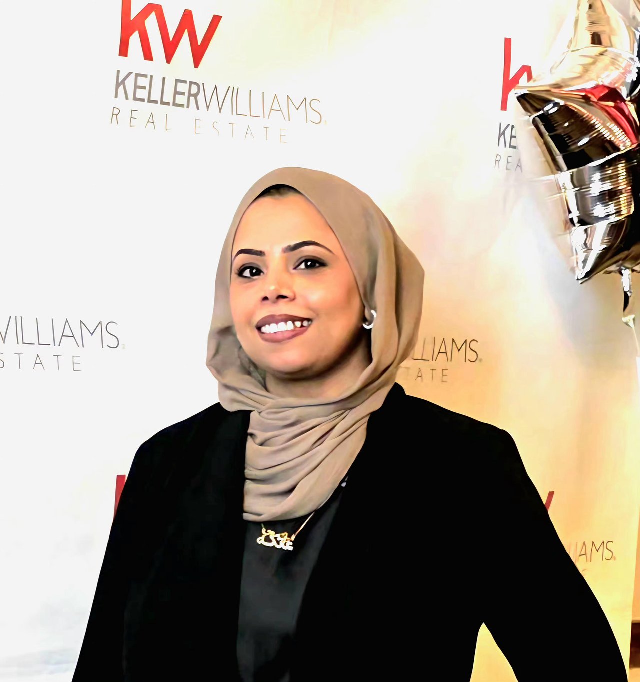 Atika Masood smiling in a beige hijab and black blazer before a Keller Williams backdrop.