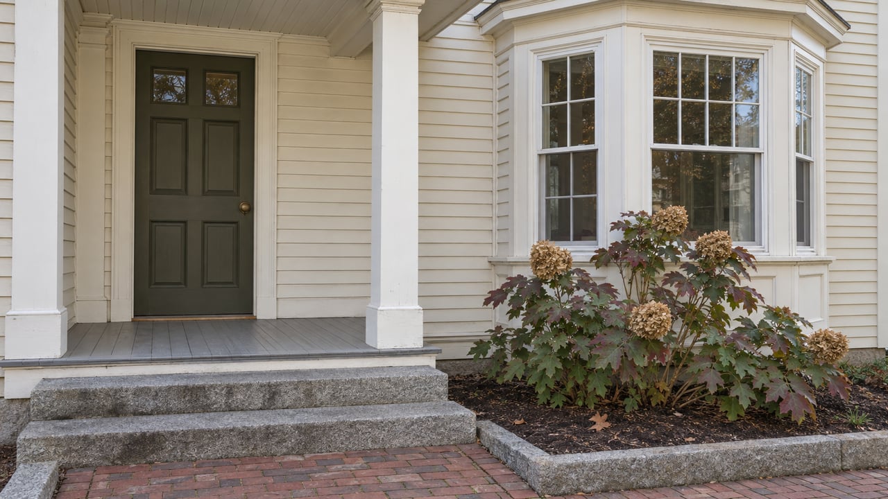 Cream clapboard home with an olive entry door, white porch column, projecting sash-window bay, granite steps and an oakleaf hydrangea.