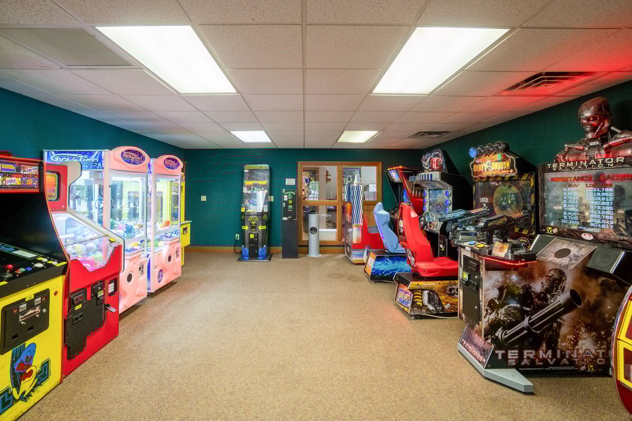 A small arcade room with various game machines, including claw machines, racing simulators, and a shooting game. The brightly lit space features fluorescent lights reflecting off green walls and carpeted floor