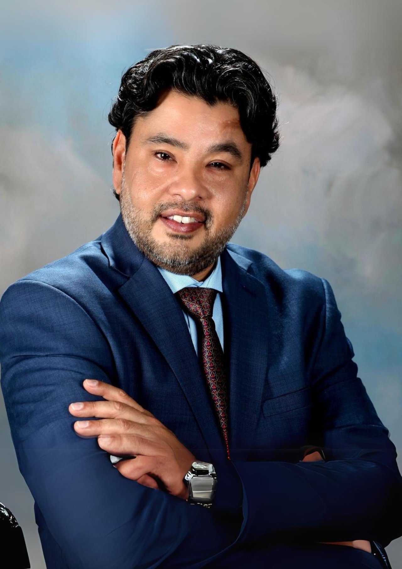 Marcus Maldonado in a navy suit and patterned tie, posing with his arms crossed.