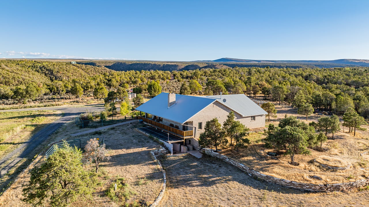 Rural stone house with metal roofs, fenced yards, and wooded hills beyond.