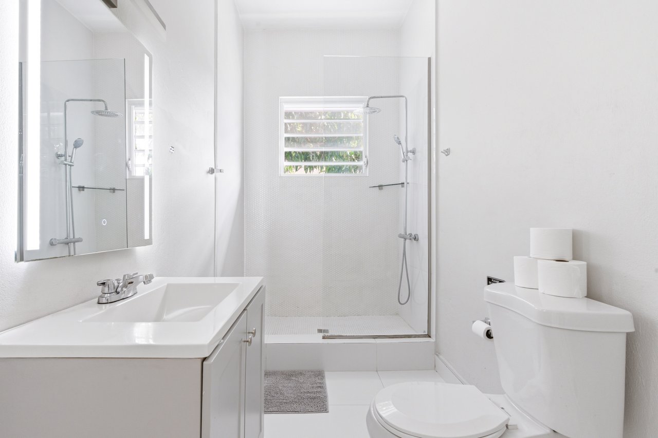 White bathroom with a glass walk-in shower, sink vanity, and window overlooking greenery.