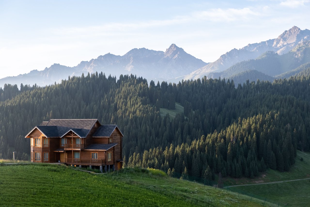 Wooden mountain cabin above an evergreen forest, with rugged peaks in the background.