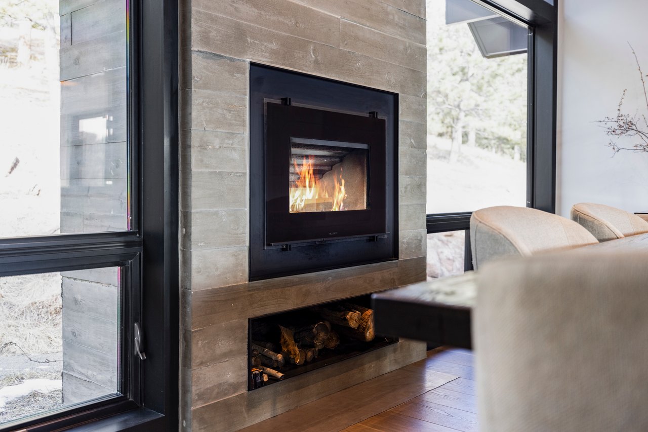 Contemporary gas fireplace with stone surround, wood storage below, and mountain views through adjacent windows