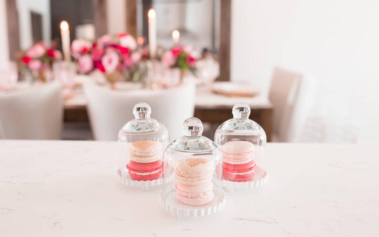 Pink and red macarons under glass cloches on a marble counter, with a candlelit floral table behind them.