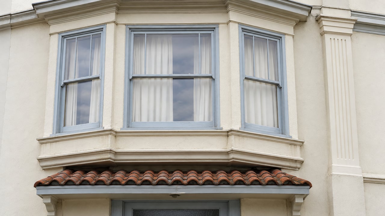 Cream stucco facade with pale blue-gray bay-window frames and a weathered terra-cotta canopy above a closed entry.