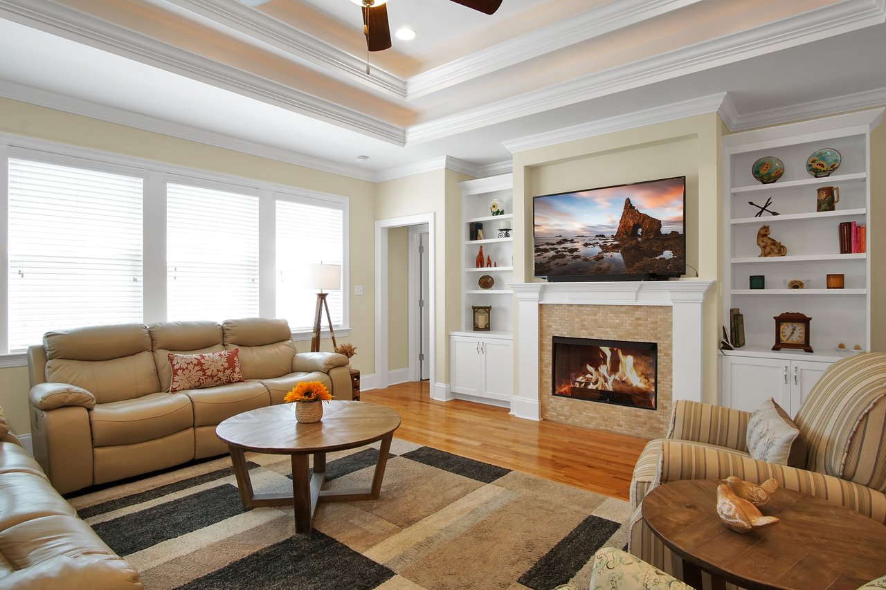 Cozy living room with beige sofas, a striped armchair, and a wooden coffee table. A fireplace with a TV above it, surrounded by built-in shelves. Warm and inviting ambiance.