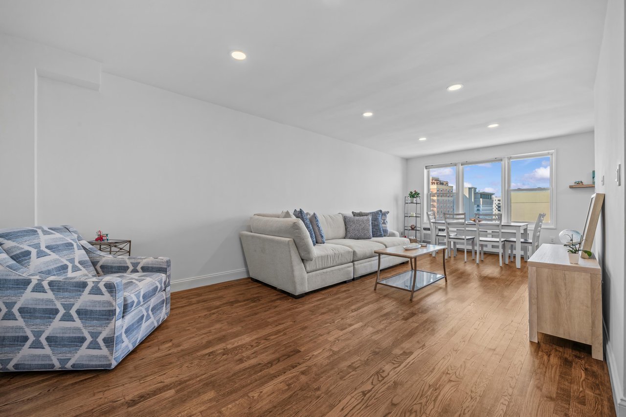 Living and dining area with hardwood floors, a gray sectional, and windows overlooking city buildings.