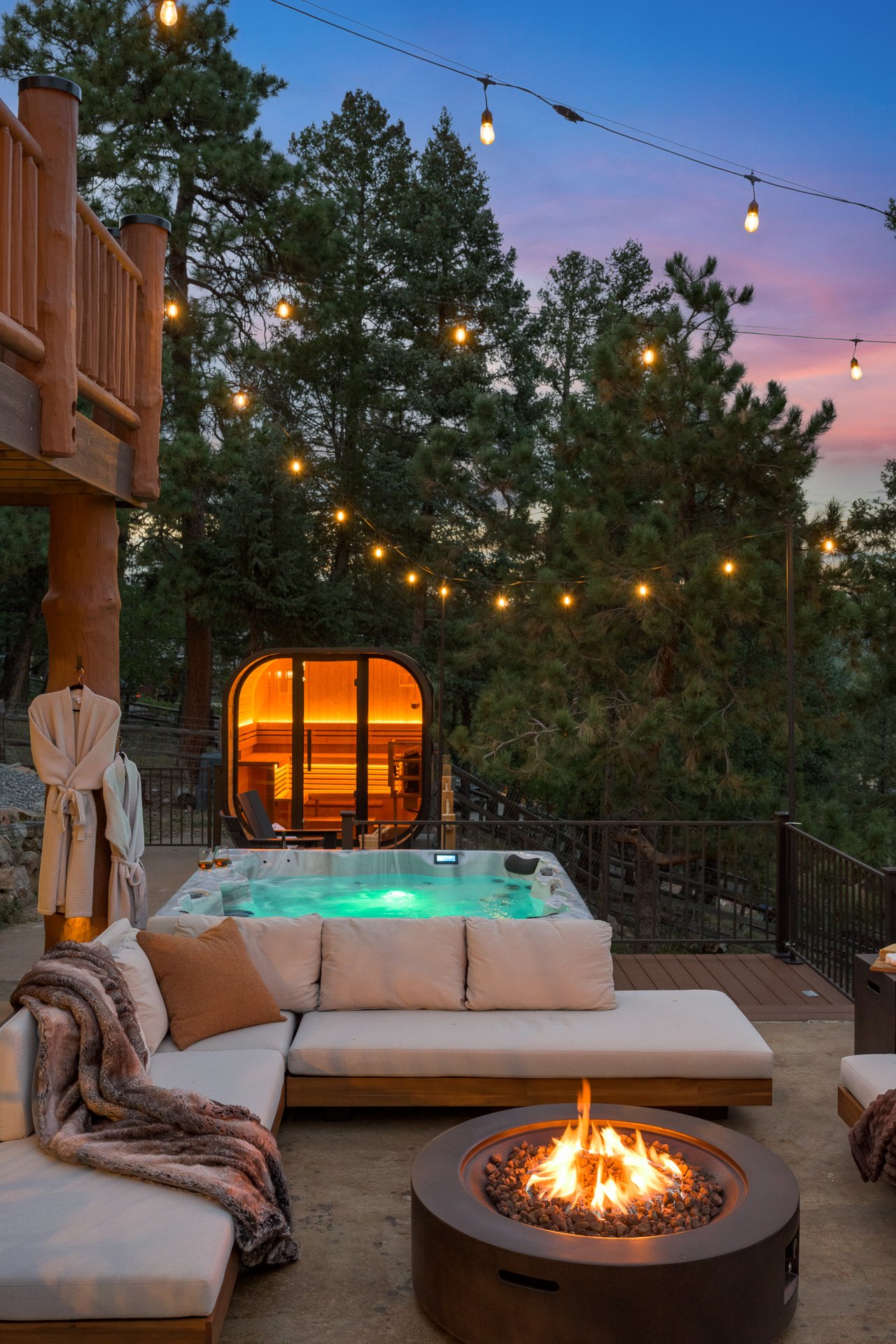 Outdoor patio with a glowing hot tub, glass-fronted sauna, cushioned seating, and fire pit under string lights.