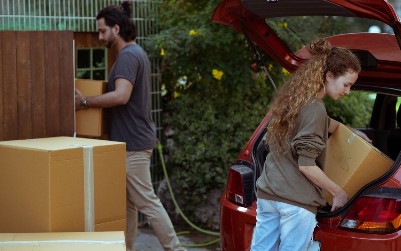 A woman loads a box into a red car as a man carries another from the house.