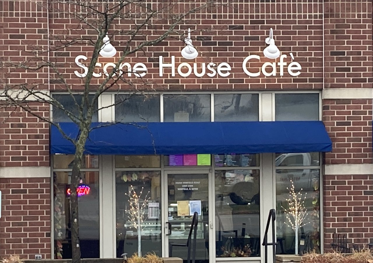 The exterior of a brick building with a sign that reads "Scone House Cafe" in large white letters mounted above a blue awning. Below the awning are glass doors and windows.