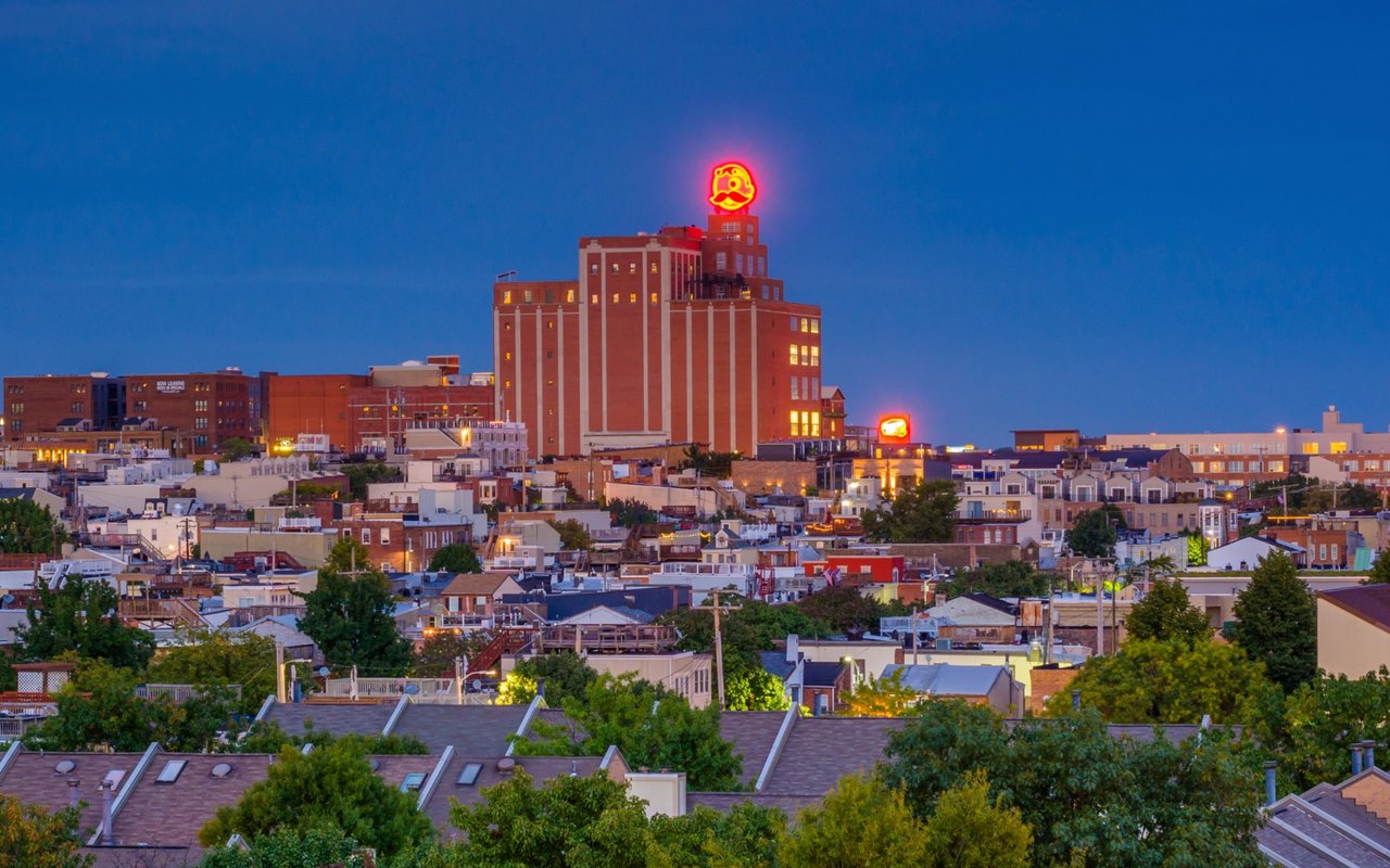 Baltimore rooftops and brewery tower topped by the glowing Natty Boh face sign at dusk.