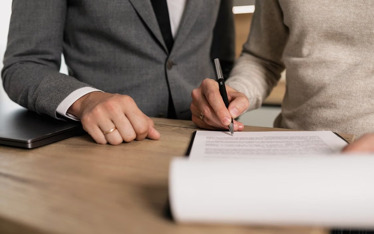 Two people review and sign paperwork at a wooden table.