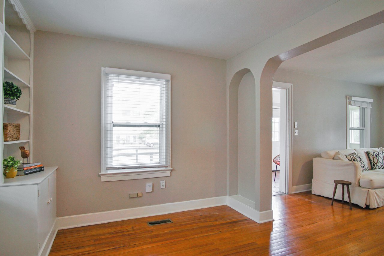 Bright living room with wooden floors, a white couch, and decorative pillows. An archway leads to another room. Shelves hold books and plants, creating a cozy feel.