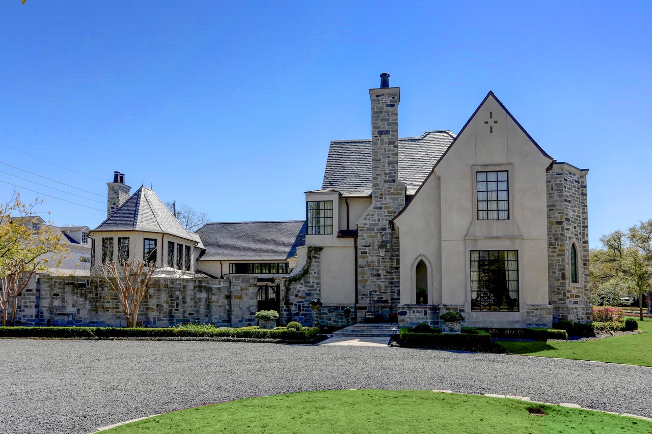 A large, French Normandy-style stone house with tall windows, a second-story balcony, a driveway, and a front lawn.
