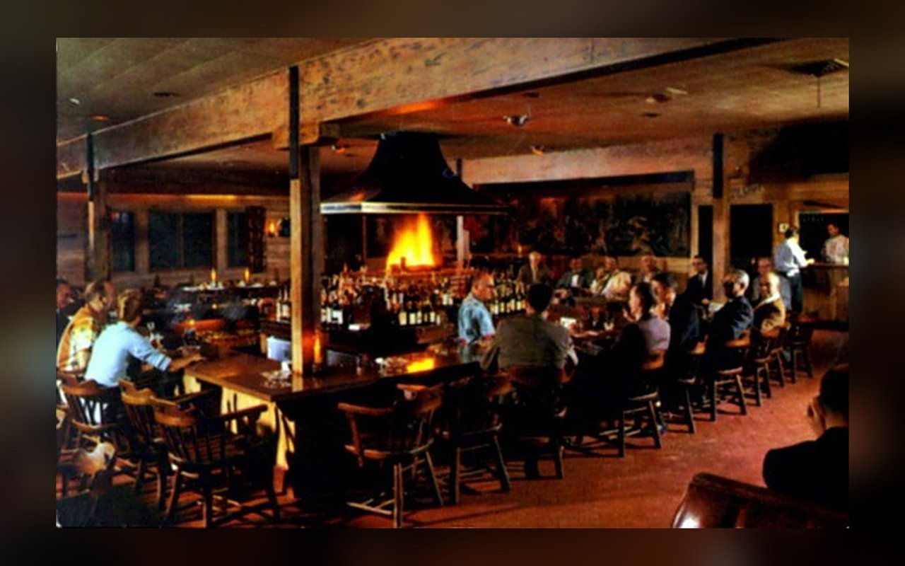 Restaurant interior with a central fireplace, long bar, exposed timber beams, and seated diners.
