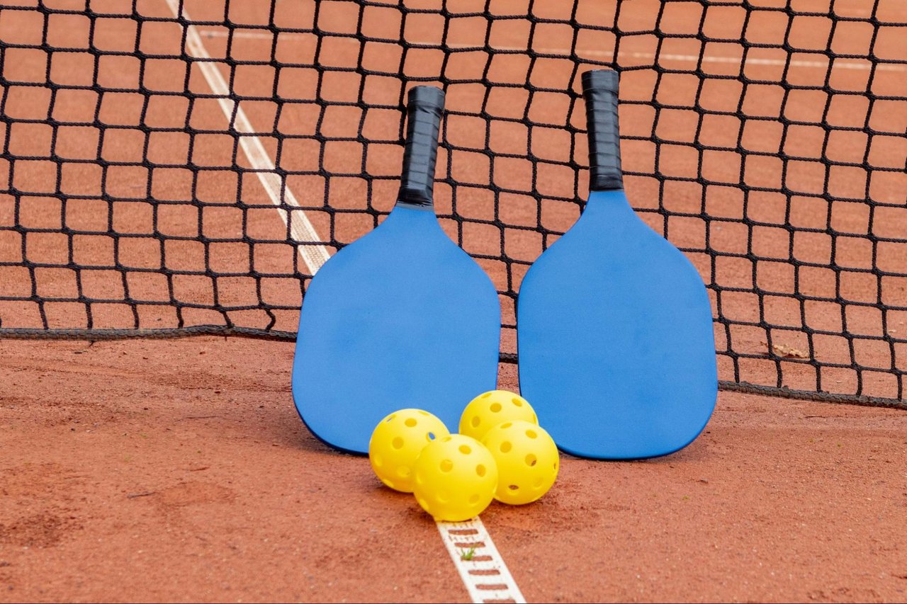 Blue pickleball paddles and yellow balls on a reddish court beside the net.