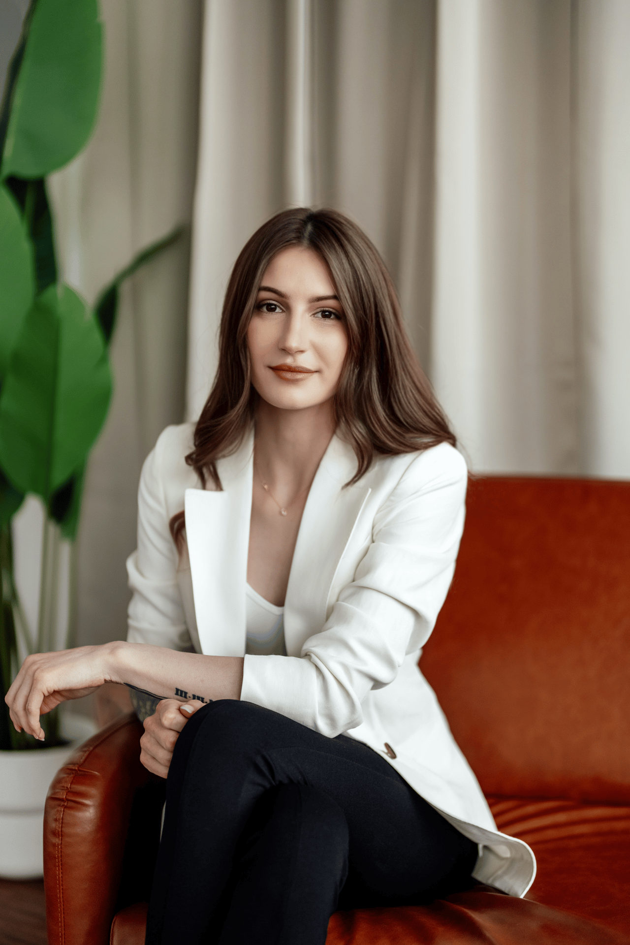 Megan Frances seated in a white blazer on a rust-colored leather chair beside a leafy plant.