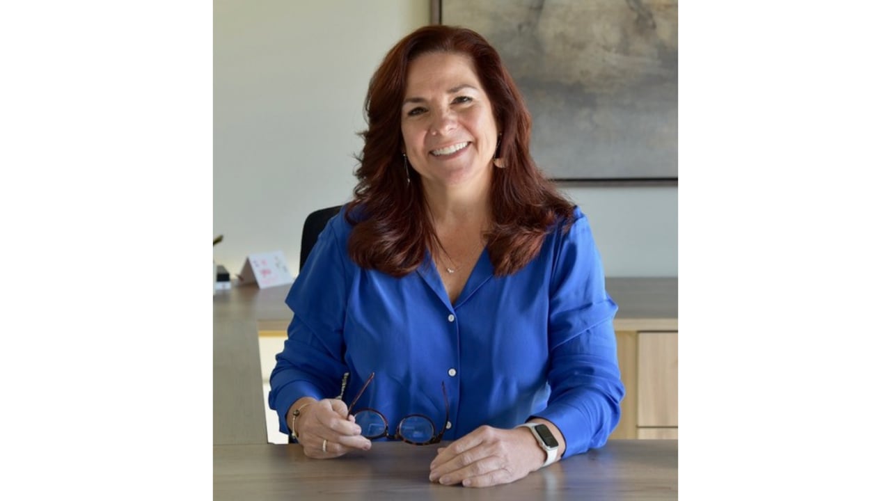 Annette Lommerin seated at a desk, smiling and holding eyeglasses.