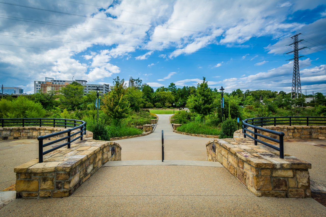 Park walkway with stone walls and railings, trees, distant buildings, and overhead power lines.