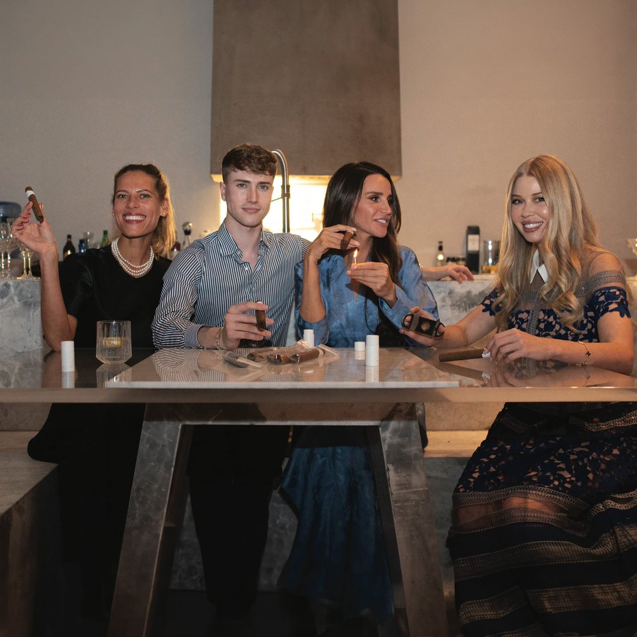 Four adults gather around a marble table, sharing cigars and drinks in a modern kitchen.
