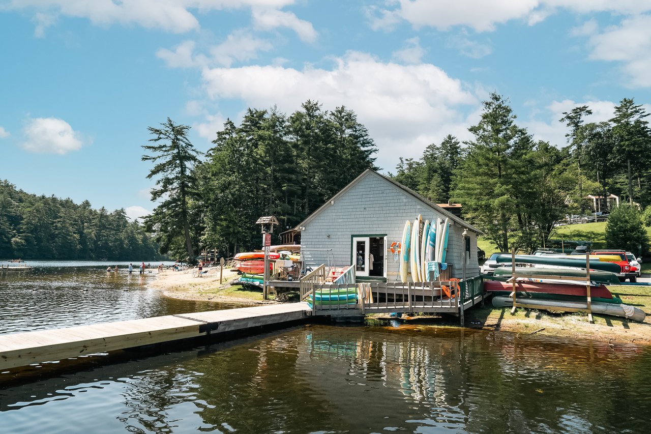 ALt=A lakeside cabin with a dock is surrounded by trees, with various kayaks and canoes stored nearby. The sky is partly cloudy and people are visible at the beach in the background. Real estate agents often highlight such serene locations to showcase potential homes for those seeking tranquility and success.