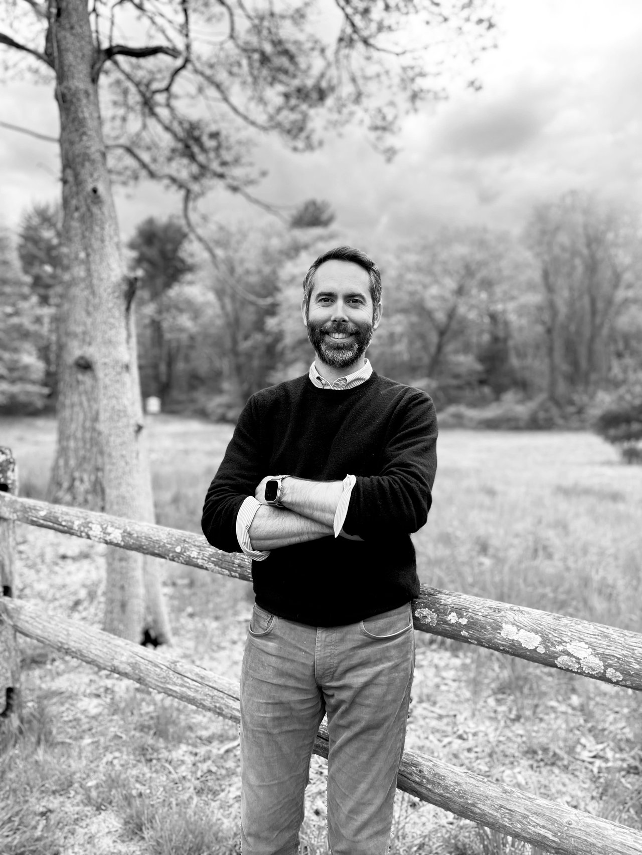 Benjamin Ginnel stands outdoors, leaning against a wooden fence with arms crossed. Trees and cloudy sky in the background. Black and white image.