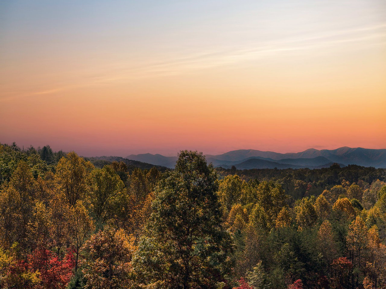 A scenic view of a North Carolina in Macon County forested landscape during sunset, with a gradient sky transitioning from orange to blue and distant mountain ranges in the background